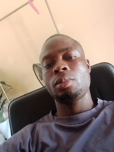 An adult male with dark skin, short dark hair, and facial stubble is depicted in a close-up, frontal portrait, looking directly at the camera with a neutral expression. He is wearing a dark, plain crew-neck t-shirt and is seated in a black office-style chair. The background consists of a light-colored wall. A portion of a circular fan grill is visible behind his right shoulder. To his left, a green leafy plant in a white pot and a stack of white objects are present. Two diagonal metal rods, one with a pink clothes clip, are visible in the upper left background. The scene is indoors, located in Okinni, Nigeria.