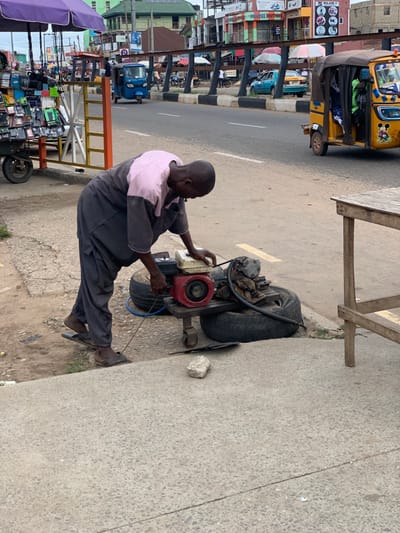 A man in a pink and gray shirt is working on a small, red engine attached to a metal frame with wheels. There are two tires on the ground next to the engine, and a blue hose is connected to it. The man is wearing sandals and is standing on a gray concrete sidewalk. In the background, there is a busy street with several vehicles, including a yellow tricycle with a Mickey Mouse sticker on the side, and several buildings. The scene is located in Ondo, Nigeria, at the coordinates 7.252 latitude and 5.199 longitude.