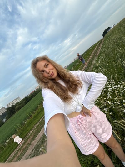 This is a medium shot, selfie-style photo taken outdoors in a field with a body of water in the distance. In the foreground, a young woman with long blonde hair is wearing a white long-sleeved shirt and pink striped shorts with red hearts. She is standing in a field of tall grass and wildflowers, with her right arm extended towards the camera. In the background, a man is walking on a dirt path, and a small child in a white dress is standing in the grass. The sky is cloudy and overcast. The location is identified as Snovitsy, Russia.