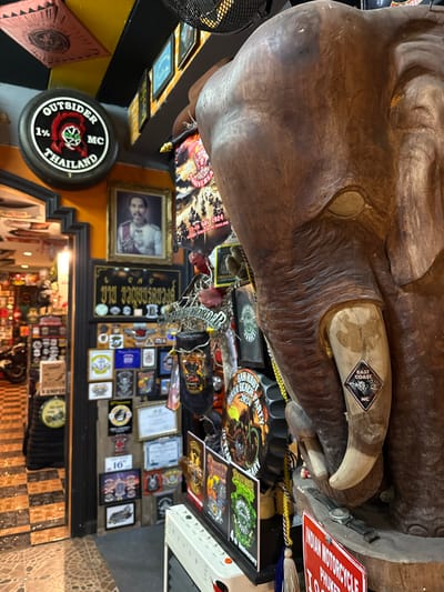 This image depicts an interior scene, likely a bar or a clubhouse, filled with motorcycle memorabilia. Dominating the foreground is a large, intricately carved wooden elephant's head with tusks. Attached to one tusk is a sticker that reads "EAST COAST MC".

The walls are densely decorated with a variety of items, including framed photographs, patches, stickers, and signs. Among the visible text are references to "OUTSIDER MC THAILAND," "INDIAN MOTORCYCLE PHUKET," "BIKER BEACH PARTY 2024," and "EAST COAST MC." There are also various club insignias and what appear to be awards or certificates. The overall atmosphere is rustic and appears to be related to a motorcycle club culture, with a focus on community and events. The lighting suggests it is likely indoors during the day or evening. The presence of "THAILAND" in multiple instances clearly indicates the location.