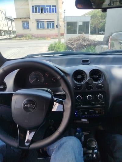 The image is a first-person interior view from the driver's seat of a Chevrolet car in Kokand City, Uzbekistan. The dashboard is visible, featuring a steering wheel with a Chevrolet logo, instrument cluster showing a mileage of 247171, air vents, and a Pioneer car stereo. Outside the car, a street scene is visible, including buildings, trees, and a small guard booth. The environment appears to be a residential or commercial area in the city. The perspective suggests the car is parked.