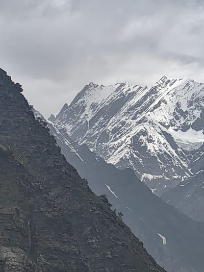 A low-angle, vertical shot captures a dramatic mountain landscape under a cloudy sky. The foreground is dominated by a dark, rocky, and rugged mountainside that fills the left half of the frame. This mountainside slopes upwards and towards the right, with visible texture and some sparse vegetation.
To the right and in the background, a majestic range of snow-capped mountains rises sharply. These peaks are jagged and angular, with significant portions covered in white snow contrasting against dark rock. The snow appears to be unevenly distributed, highlighting the steep slopes and deep crevices. Patches of snow are also visible on the lower, darker mountain slopes.
The sky above is overcast with diffused light, creating a muted and somber atmosphere. The overall impression is one of a remote and imposing natural environment. The location is identified as Unknown, India.