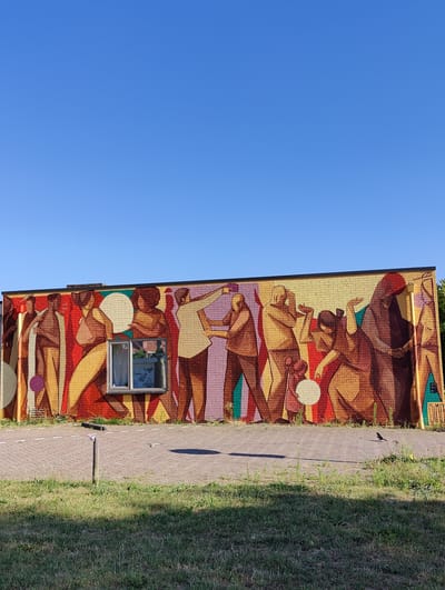 The image depicts a mural on a brick building, featuring stylized human figures in warm, earthy tones against a clear blue sky. The mural, located in Breda, Netherlands, spans a significant portion of the building's facade. In the foreground, a paved walkway with painted lines separates a strip of green grass from the building. A small bird is visible on the walkway, and a wooden post with a red and white marker stands in the grass. A window is integrated into the mural, reflecting the surrounding environment. The overall scene is one of urban art meeting everyday life under a bright sky.