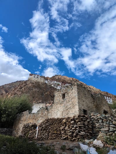 This image captures a dramatic scene of a monastery built precariously into a rocky cliff face. The monastery's white buildings with red trim stand out against the rough, brown stone of the mountain. The structures appear to be multi-storied and cling to the sheer rock, suggesting a remarkable feat of engineering and a deep connection to the natural environment.
Below the main monastery, a section of the cliff is reinforced with a wall constructed of numerous stones, creating a textured and robust barrier. Some of the stones are quite large, while others are smaller and more rounded, indicating a careful selection and placement. Green foliage, including trees and bushes, grows around the base of the cliff and along the edges of the constructed areas, adding a touch of life to the arid landscape.
The sky above is a vibrant blue, filled with fluffy white clouds that are dramatically illuminated by sunlight, suggesting a bright and clear day. The angle of the light indicates it could be late morning or early afternoon. The overall impression is one of resilience, devotion, and a harmonious coexistence between human habitation and the rugged terrain.
The location is identified as Yulang, India, which is known for its mountainous landscape and spiritual sites. There is no visible text within the image.