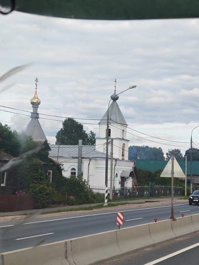 The image shows a street scene in Maryino, Russia, with a white church with a golden dome on the left. In the foreground, there is a road with a concrete barrier. A black car is driving on the road. The sky is cloudy. There are also some trees and a fence in the background.