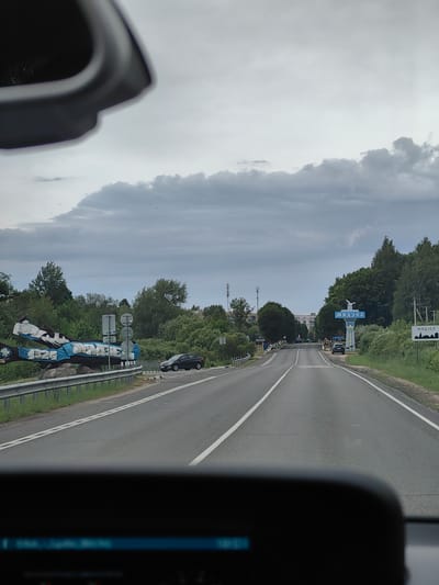 The image captures a view from inside a vehicle, looking forward along a paved road in the city of Мядзельскі сельскі Савет, Belarus. The sky is overcast with gray clouds. On the left side of the road, there is a large, sculptural art installation resembling a stylized aircraft, painted blue and white with lettering. Adjacent to this is a metal guardrail and a grassy embankment. A black car is parked on the shoulder to the right of the art installation.
Further down the road, on the right side, a sign with blue lettering that reads "МЯДЗЕЛ" (Myadzel) and a white silhouette of a bird on top is visible. This sign is mounted on a decorative pillar. Another smaller sign with a similar silhouette and the word "МЯДЗЕL" is also present. A few people are walking on the sidewalk near the "МЯДЗЕЛ" sign. Trees and buildings line the sides of the road. In the foreground, the dashboard of the vehicle is partially visible, showing a dark, reflective surface. Several birds are seen flying in the sky and above the trees.