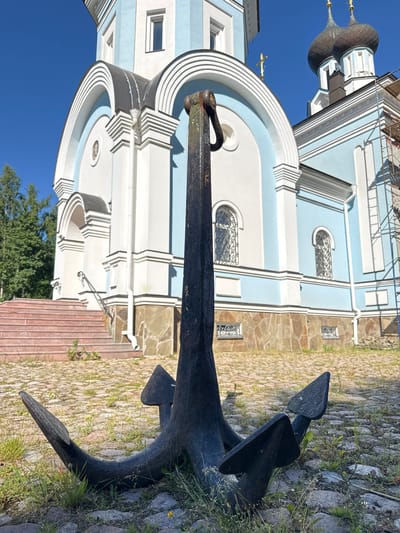 The image captures an ancient anchor placed in the foreground on a cobblestone path, overgrown with grass. In the background, a pale blue and white church with domed roofs stands under a clear blue sky. To the left of the church, a set of reddish-brown stairs leads up to an entrance. Scaffolding is visible to the right of the church, indicating ongoing construction or renovation. The overall setting appears to be the village of St. Ozerki in Russia.