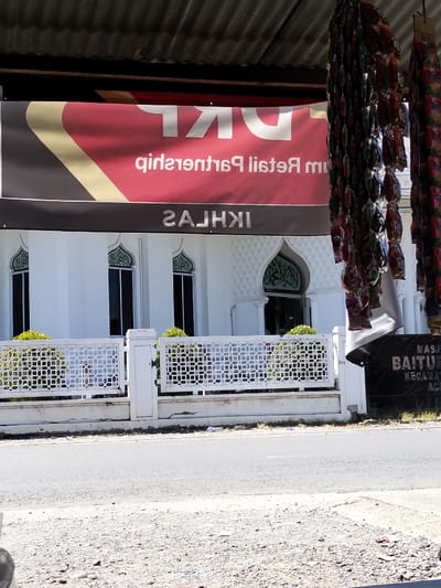A mosque with white walls and an arched entrance stands behind a white fence. A sign for "Retail Partnership" is displayed above. To the right, hanging strands of snacks are visible. A black sign for Masjid Baiturrahim is also present. The scene is viewed from across a road.
