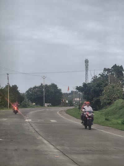 The image captures a cloudy day scene on a rural road, likely in Padli, India. A paved road curves through a landscape dotted with trees and vegetation. Two motorcycles are visible on the road. The first motorcycle, closer to the foreground, is being ridden by two people and is moving away from the viewer, with the rear brake light illuminated. The second motorcycle is further ahead and also moving away. In the background, a tall cell tower stands prominently against the overcast sky. Other elements include utility poles with wires, some traffic signs, and a flag on a pole. The overall impression is one of a typical rural roadside scene under a gray sky.