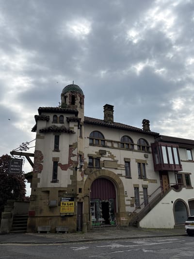 This is an exterior shot of The Ruskin Rooms building in Knutsford, United Kingdom, taken on a cloudy day. The building is constructed with a mix of white and tan stone, and its architecture features a prominent tower with a green, domed roof. The building exhibits both architectural details and signs of age, with brickwork exposed in some areas. A "To Let" sign is posted on the side of the building, advertising office space for rent. A sign with the name "The Ruskin Rooms" hangs from a post. Part of a car can be seen on the street.