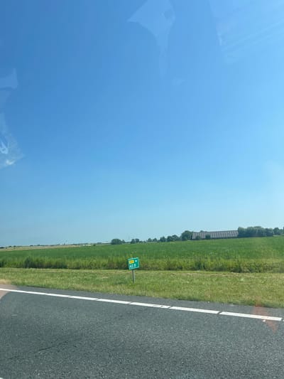 The image shows a bright, clear blue sky over a grassy field next to a road. The scene appears to be a rural landscape in Marsum, Netherlands, during the daytime. A long, multi-story building with many windows is visible in the distance, partially obscured by trees. A green road sign with white text is posted along the side of the road, indicating "N31 Re 42.8". The road itself is dark asphalt with a white line marking the lane. The sun is shining brightly, casting reflections on the sky. There are no people visible in the image, and the overall impression is one of a quiet, sunny day.