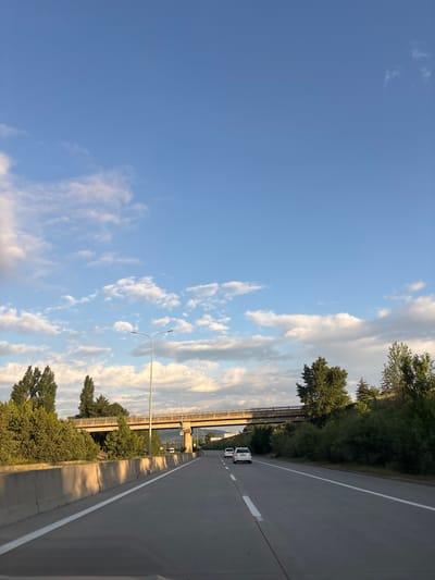 The image shows a highway in the city of Berbuki, Georgia, under a bright blue sky with scattered white clouds. A concrete barrier runs along the left side of the road, casting long shadows. Trees and foliage line the sides of the highway, with a bridge crossing overhead in the distance. Two white cars are visible on the road, traveling in the same direction as the viewer. The scene appears to be captured during daylight hours, likely late afternoon, given the angle of the sun and the shadows.