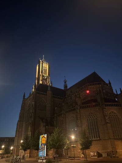 The image shows a large, historic brick church illuminated at night against a dark blue sky. The church has a tall, prominent tower with ornate detailing and a spire. The building's facade features gothic-style windows and architectural embellishments. A bright red light is visible on a section of the church's roof. In the foreground, there are several trees, streetlights, and bicycles parked. A signpost with various pictograms and text, including a bicycle symbol and pedestrian crossing, is also present. The overall scene suggests a nighttime view of a city square or street in front of the church. The text on the signposts indicates the location is Arnhem, Netherlands, and references "zero-emissie" which translates to "zero emission," likely relating to a city initiative.