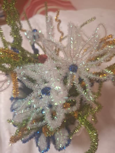 The image displays a close-up, slightly out-of-focus view of a festive arrangement, possibly a small Christmas tree or a decorative centerpiece. The arrangement is composed of various metallic pipe cleaners and tinsel in colors including white, green, gold, and blue. Some sections are shaped like stars or snowflakes, while others resemble evergreen branches. Small, spherical blue ornaments are interspersed throughout the arrangement, along with a few red accents. The background is soft and out of focus, suggesting a fabric surface, possibly a tablecloth or blanket, with hints of a striped pattern in the upper portion of the image. There is no visible indication of the city of Mıñbaev, Kazakhstan, or any specific environmental details that would place the scene there. The lighting appears to be artificial, casting soft highlights on the metallic elements. The overall impression is one of holiday decoration.