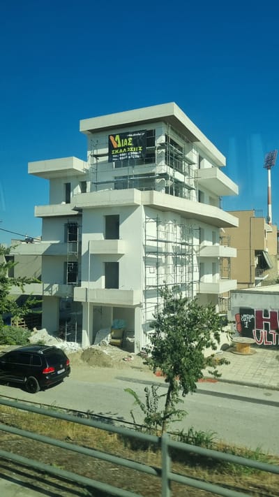 The image shows a multi-story building under construction in Larissa, Greece. The building has a white exterior and is covered in scaffolding. A black SUV is parked on the street in front of the building. The sky is clear and blue. There is a banner on the building that reads "MIAS" in Greek and includes a website address and phone numbers. There is also graffiti on a wall in the background that says "LARISSA GATE 1 ULTR...". The scene appears to be an outdoor urban environment with some greenery.