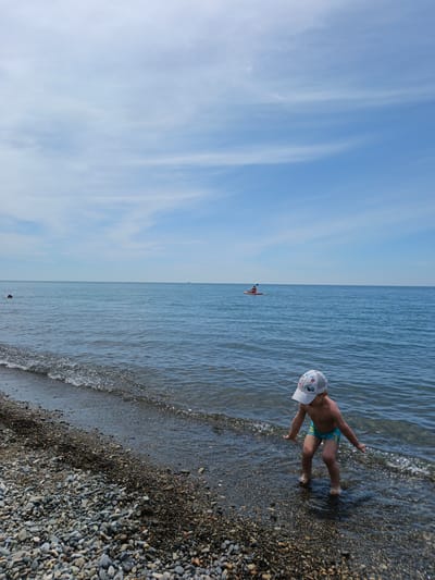 This vertical photograph captures a daytime scene at a pebble beach in Krinitsa, Russia. In the foreground, a young child wearing light blue swim trunks and a white baseball cap with colorful designs stands in the very shallow water at the edge of the dark, multi-colored pebble beach. The child is bent forward with arms slightly spread, facing the water, with small waves breaking around their bare feet. In the middle ground, a person in an orange kayak paddles leftward across the calm, blue-grey sea, creating a subtle wake. Further to the left, a swimmer's head and shoulders are partially visible above the water. The upper portion of the image displays a clear blue sky with sparse, wispy white clouds. The horizon line is flat, separating the calm sea from the sky.