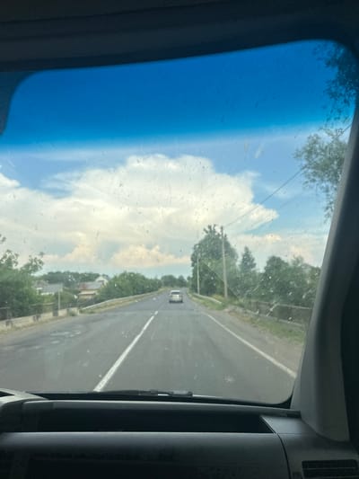 The media file depicts a view from inside a vehicle, looking out through the windshield. The scene is dominated by a bright blue sky with scattered white clouds, suggesting a daytime setting. A paved road stretches into the distance, marked with a white line. On either side of the road, lush green trees line the landscape, with some hints of houses visible amongst them. A single white vehicle is seen on the road ahead. The windshield itself appears somewhat smudged or dirty, with small marks scattered across its surface. The overall impression is one of a scenic drive through a rural or suburban area under pleasant weather conditions. The image orientation is tilted, as if the camera was held at an angle. There is no visible text in the image.