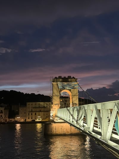 A vertical, low-angle exterior shot captures a bridge over a body of water at twilight in Vienne, France. The sky is a deep, dusky purple with streaks of pink and blue. The bridge, constructed of light-colored metal with visible rivets and beams, stretches across the frame from right to left. A stone tower, illuminated by warm lights, stands on the left side of the bridge, supporting the suspension cables. Blue and white banners are draped across the top of the tower.

In the background, buildings with illuminated windows are visible along a waterfront, with a dark, tree-covered hill rising behind them. The water reflects the lights from the bridge and buildings, creating shimmering patterns. The scene is devoid of people.