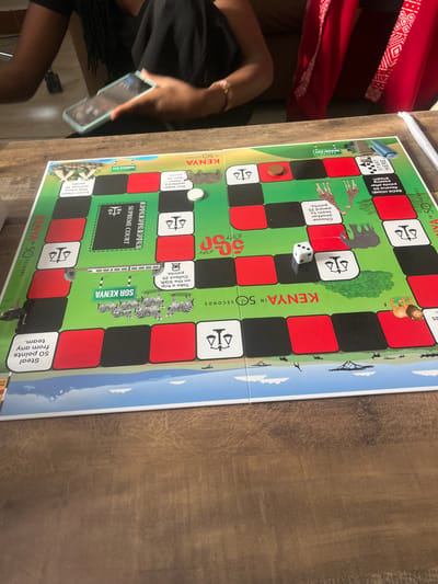 This image depicts an indoor scene in Ongata Rongai ward, Kenya, featuring a board game spread across a light brown, textured wooden table. The central object is a "Kenya in 50 Seconds" board game, characterized by a path of alternating red and black squares on a green background. The board includes illustrations of Kenyan wildlife such as zebras, giraffes, elephants, and a lion, alongside designated areas labeled "Supreme Court," "SGR Kenya," and "Nairobi City." Visible game components on the board include a white circular token, a brown circular token, and a white six-sided die displaying the numeral "2" on its uppermost face. Two individuals are partially visible seated around the table. On the left, a person with dark skin and visible braided hair, wearing a black top, holds a smartphone with a light blue case in their left hand. A multi-colored bracelet is present on their right wrist. On the right side of the frame, another individual's lower torso is visible, clad in a vibrant red garment adorned with white geometric patterns.