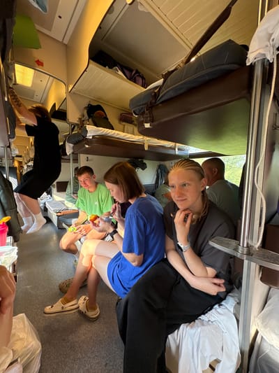 The image depicts the interior of a train compartment, likely on the Trans-Siberian Railway, given the context of Sovkhoznyi, Russia. Four young people are visible. On the left, a person in a black t-shirt and shorts is hanging from a metal bar near the ceiling. In the middle, a young man in a bright green t-shirt sits, holding a piece of fruit and looking towards the person on his left, who is a young woman in a blue t-shirt eating with a utensil. To the right, another young woman with braided hair is seated and looking forward, wearing a dark t-shirt and a smartwatch. The compartment features bunk beds, luggage racks, and a window showing greenery outside. The overall atmosphere suggests a journey in progress.