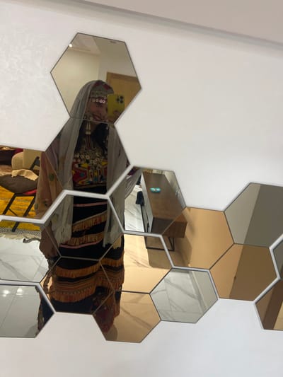 A collection of gold and mirrored hexagonal tiles adorns a white wall, reflecting a room interior. A woman in traditional attire is seen in the reflection, taking a selfie with a phone. The room features a wooden desk, a chair, and a hint of yellow and red fabric. The mosaic creates a fragmented view of the scene, blending the room's elements with the woman's self-portrait.