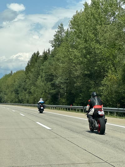 Two motorcyclists are riding on a highway in Tragail, Austria. The motorcyclist in the foreground is wearing a black and red leather racing suit and a black helmet. The motorcycle has a red rear wheel and a license plate that reads "W 7 SSN". The motorcyclist in the background is wearing a blue and white suit and a white helmet. The motorcycle is black. The highway is paved and has multiple lanes. On the right side of the highway, there is a metal guardrail and a dense line of trees. The sky is cloudy with patches of blue.