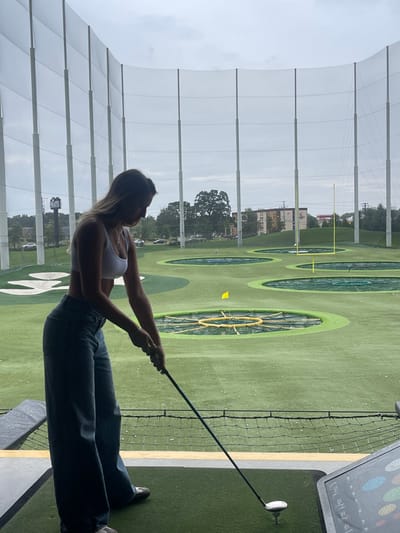 A female individual is depicted in profile, positioned within a golf bay at a Topgolf facility in Oxon Hill, Maryland, United States. She has light-colored hair and wears a white athletic bra top and blue wide-leg jeans. Her hands grip a golf driver, poised over a white golf ball resting on a white tee on a green artificial turf mat, indicating an imminent swing. To the right of the mat, a digital interface panel is visible. The expansive outdoor driving range in the background features multiple circular targets of varying sizes on artificial turf, with one target marked by a yellow flag and another containing a yellow American football goalpost. Tall white poles connected by a white protective netting enclose the range. Beyond the netting, trees, multi-story buildings, and parked vehicles are visible under an overcast sky.
