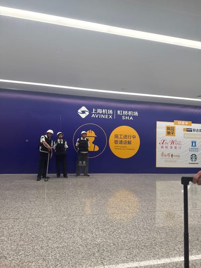 The image depicts a section of a large, brightly lit indoor space, likely an airport terminal or a similar public transit hub in Changning District, China. A prominent blue wall serves as a backdrop for several visual elements. On the left side of the wall, three security personnel are positioned. The person on the far left is wearing a white hard hat, a black vest over a white shirt, and dark pants, and is holding a long, metallic pole with a grey handle. The person in the middle also wears a white hard hat, a black vest over a white shirt, and dark pants. The person on the right wears a similar uniform with a white hard hat and a black vest. Next to the security personnel is a large circular graphic in yellow and orange. This graphic contains Chinese text that appears to read "施工进行中 敬请谅解" which translates to "Work in progress, thank you for your understanding." to the right of this graphic is a sign with multiple logos. The logos include "剪花娘子", "united money", "联合货", "BeanStar比星", "Xin Wang Restaurant", "新旺茶餐厅", "康师傅牛肉面", and "Starbucks". The floor is paved with speckled grey tiles. In the foreground, a black suitcase handle is visible on the right edge of the frame, suggesting that someone is holding a suitcase. The overall scene conveys a sense of ongoing construction or renovation within a modern, well-maintained public area.