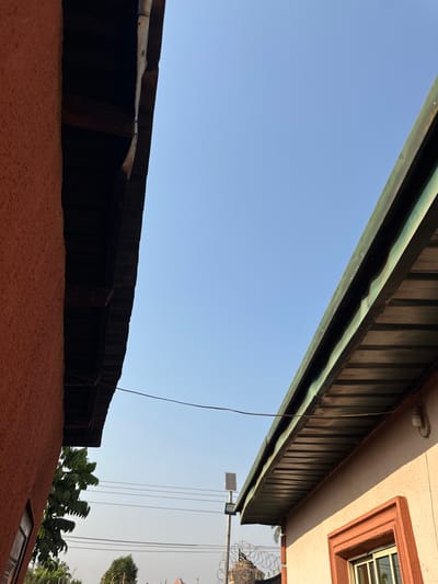This image, taken in Agbogugu, Nigeria, depicts a sky view between two building structures. On the left, a building with an orange-red textured stucco wall is visible, with its dark brown wooden eaves extending into the frame from the top left. On the right, a building with a light beige wall features a green corrugated metal roof and eaves, also extending into the frame from the top right. A window with reddish-brown trim and an external light fixture are visible on the lower right building. A thin, dark brown wire spans horizontally across the middle of the frame, connecting the two sides. The sky above is clear light blue, transitioning to a lighter, hazy shade towards the horizon. In the mid-ground, a tall metal pole with a solar panel and an associated light fixture is prominent. Below the solar light, barbed or concertina wire is visible on top of a structure. Multiple horizontal utility wires are also present in the mid-distance. Green foliage from trees or bushes is visible on the far left and in the distant background. No individuals or direct interactions are observable.
