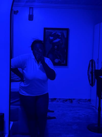 A person wearing a white t-shirt is positioned in a room with a blue hue. The person's hand is partially covering their face. A framed picture hangs on the wall behind them. A ceiling fan is visible above. The scene appears to be indoors. There is no evidence that the image was taken in Rumuigbo, Nigeria.