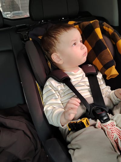 A young child, appearing to be a baby or toddler with light brown hair, is seated in a dark car seat within a vehicle. The child is looking towards the upper right with a calm, observant, or slightly pensive expression. They are dressed in a light-colored, long-sleeved shirt with faint horizontal stripes and light grey pants. A yellow and black plaid blanket or jacket is draped over the top of the car seat, behind the child's head.

The car seat features a black 5-point harness system with dark brown shoulder pads, one of which has a partially visible "bo" logo. A black buckle with a red release button secures the harness. On the child's lap, there is a small yellow toy dump truck and a white fabric item with red polka dots or patterns. To the child's left, a dark-colored jacket or blanket is crumpled on the car's back seat.

The interior of the car is visible, including the dark leather headrests of the front seats. Through the blurred rear window, other vehicles and an overcast daytime sky are discernible. A yellow "AIRBAG" label is visible on the side of the car seat. The scene depicts a child traveling in a car, in what is contextually specified as Novoe Mart'yanovo, Russia.
