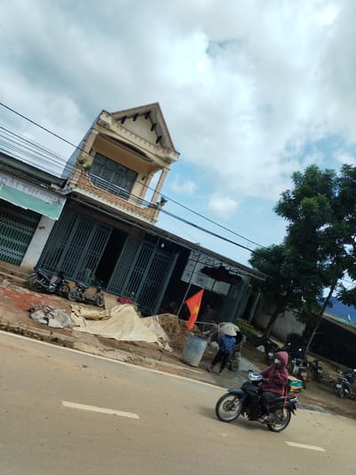 The image shows a street scene in Pờ Y, Vietnam. A two-story building with a faded yellow facade is angled in the frame. The ground floor features large, closed metal doors, and several motorbikes are parked nearby. Construction materials and equipment are scattered on the adjacent sidewalk, including a Vietnamese flag.

A person is operating a concrete mixer behind a blue barrel. Another person is on a black motorbike, driving along the road. A tree is in the background. The sky is partly cloudy.