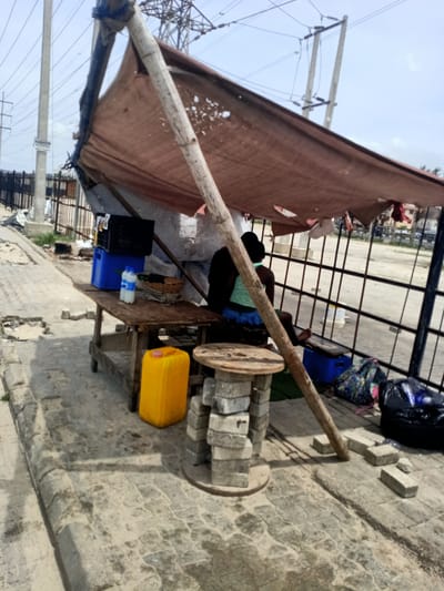 The image captures an informal roadside stall in Eputu Town, Nigeria, under a clear, bright daytime sky. A person, seen from behind, sits on a low stool beneath a makeshift shelter constructed from rough wooden poles and a torn, brown fabric tarp, which provides shade.

The stall features a rustic wooden table holding a stack of blue and black crates, a white plastic bottle with the word "BIGI" visible on its label, and a woven basket. A yellow plastic jerrycan rests on the ground beside the table. Another improvised piece of furniture, a round wooden spool supported by stacked bricks, serves as a side table or display surface.

The ground is paved with interlocking concrete blocks, appearing somewhat worn and dirty. In the mid-ground, a black metal fence or gate runs horizontally. The background reveals utility poles with power lines stretching across the sky, and a large metal electricity pylon is visible further in the distance. A white bucket can be seen through the fence.

On a utility pole to the left, a sign is attached, displaying the text "FASHION SCHOOL" and a phone number "08037704027." The scene suggests a small-scale vendor operating or resting at their humble stall. A colorful backpack and a black plastic bag are visible on the ground near the seated individual. The overall setting depicts a typical informal commercial setup in a semi-urban or rural Nigerian environment.