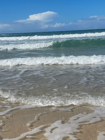 The image depicts a view of the ocean from a beach on the Gold Coast, Australia. Multiple waves are rolling towards the shore, with white foam cresting on their tops. The water is a clear turquoise green. The sky is blue with scattered white clouds. The sand in the foreground is wet and appears to be shallow water lapping at the shore. There are no people or man-made objects visible in the frame.