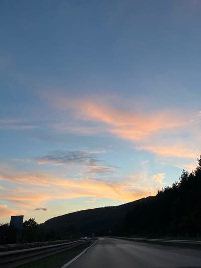 The image depicts a scenic view from a highway in the city of Churek, Bulgaria, during sunset. The sky is a blend of blues and soft pinks, illuminated by the setting sun. Wispy clouds are scattered across the horizon, catching the warm hues of twilight. A dark, tree-covered hill slopes down to the right of the highway, its silhouette stark against the colorful sky. The highway itself stretches forward, curving gently to the left. A metal guardrail runs along the side of the road, separating it from the shoulder. On the left side of the highway, a dark, rectangular structure, possibly a sign or part of an infrastructure, is visible amongst trees. The overall atmosphere is tranquil and serene, characteristic of a peaceful evening drive.