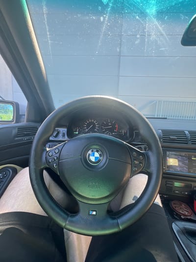 The image shows the interior of a BMW car, looking forward from the driver's seat. The steering wheel is prominently in the center, with a BMW logo on it. Various buttons and controls are visible on the steering wheel and the dashboard, including a digital display screen that shows what appears to be a map or navigation. The car's instrument cluster is visible behind the steering wheel, showing the speedometer and tachometer. The driver's legs are visible in the lower part of the image, with one leg wearing black pants and the other exposed, showing skin with visible hair. The car's interior suggests it is a relatively older model. The windshield is visible at the top, with sunlight streaming through, creating reflections and glare. The exterior environment is suggested by the reflection in the side mirror, showing trees and a building with a corrugated metal facade, possibly a garage or industrial building. The scene appears to be set in a sunny outdoor location. The exact location of Koziegłowy, Poland, cannot be determined from the image alone.