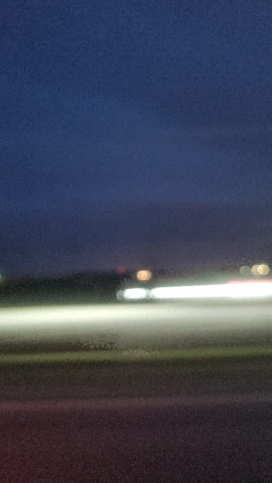 The image displays a highly blurred, low-light scene, consistent with a long-exposure photograph captured at night or late dusk. The entire frame exhibits significant motion blur and an absence of sharp detail.
The upper approximately 50% of the image is dominated by a uniform dark blue-indigo sky, devoid of any discernible clouds or celestial bodies. Below this, a faint, dark horizontal band marks what appears to be a distant horizon or treeline.
In the mid-ground, above the prominent light streaks, several indistinct, out-of-focus light sources (bokeh) are visible against the darker backdrop. These include a distinct circular golden-amber light positioned slightly right of the center and a smaller, fainter golden light further to the right.
The lower half of the image is characterized by horizontal streaks of light. Two prominent, parallel, bright white light streaks extend from the left to the right across the mid-bottom section, indicative of vehicle headlights or taillights in motion during a long exposure. Below these white streaks, a broader, diffuse horizontal band of light grey and faint greenish hue is present, likely an illuminated surface or reflected light. The lowest portion of the frame shows a dark, grainy surface, consistent with a road, displaying subtle hints of reddish-brown and dark grey.
No discernible individuals, specific vehicles, or identifiable structures are present. The precise location within Norrköping, Sweden, cannot be determined from the visual information provided in this image.