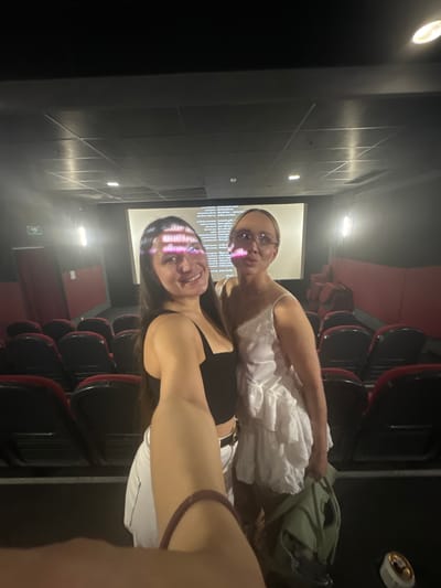 Two women are taking a selfie in a movie theater. The theater is dark with red seats. The movie screen is visible in the background, showing the credits. One woman is wearing a white dress and glasses, while the other is wearing a black top and white pants. They are both smiling. A green bag is on the seat next to the first woman. A white container is above them.
