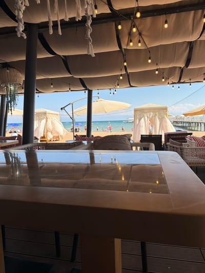 This is a medium shot from inside a covered outdoor seating area overlooking a beach scene. In the foreground, a reflective surface, likely a table, shows reflections of string lights and the interior of the structure. Beyond the table, there are outdoor seating elements, including chairs with red and white striped cushions and woven baskets. In the midground, a sandy beach is visible with several white cabanas, beach umbrellas, and numerous people scattered across the sand and in the water. A wooden pier extends into the lake on the right side of the image. The background features a body of water, likely Issyk-Kul Lake, and a clear blue sky. The overall impression is a beachfront resort or cafe setting in Sary-Oy, Kyrgyzstan.