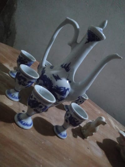 The image displays a collection of traditional blue and white ceramic ware, likely a tea or wine set, arranged on a reddish-brown wooden surface. The central object is an elegant ceramic pitcher or teapot featuring an elongated, upward-curving spout and a distinct handle. Its white porcelain body is intricately decorated with blue patterns, possibly depicting dragons or mythical creatures, in a classic blue-and-white style.

Surrounding the pitcher are several matching small, goblet-shaped cups. Each cup has a narrow stem and a flared base, adorned with blue patterns that complement the pitcher's design. At least five cups are visible in the frame, casting prominent shadows. A small, light-colored ceramic figurine, possibly resembling a rabbit or duck, is also present on the wooden surface towards the upper right of the image, contributing to the shadows.

The scene is dimly lit, suggesting either low ambient light or nighttime, which emphasizes the contrast between the objects and their elongated shadows on the wooden surface. The background consists of a dark grey wall. The objects appear to be stationary, either displayed or awaiting use in an indoor setting. No discernible text is visible on any of the items.