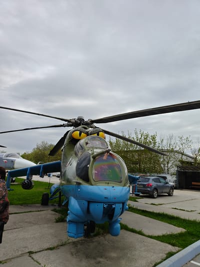 The image depicts a Mil Mi-24 Hind attack helicopter displayed outdoors, presumably as a static exhibit. It is located in Arkhangel’skoye, Russia, as evidenced by the license plate on the gray Nissan parked in the background. The helicopter is painted in blue and green camouflage with stylized yellow "eyes" on the nose, possibly added for amusement. The rotor blades are black. The helicopter is situated on concrete slabs with patches of grass growing between them. Another aircraft, possibly a fighter jet, is partially visible in the background on the left. The sky is overcast. There is a person partially visible in the lower left corner of the image. There is a wooden shed and a few trees in the background behind the gray Nissan. A red ribbon is placed inside the cockpit.