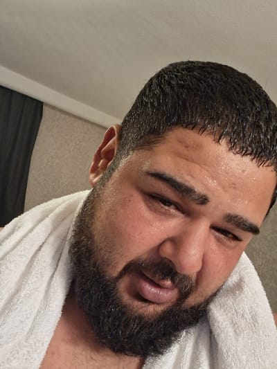 A close-up, overhead shot shows a person with dark hair and a beard, wearing a white towel draped around their neck. The person's face is sweaty, with visible perspiration on their forehead, cheeks, and the bridge of their nose. Their eyes are partially closed, and their lips are slightly parted. The background consists of a textured wall and a dark curtain. The lighting appears to be from above, casting a soft glow on the scene. The location is not identifiable as Thüngersheim, Germany.