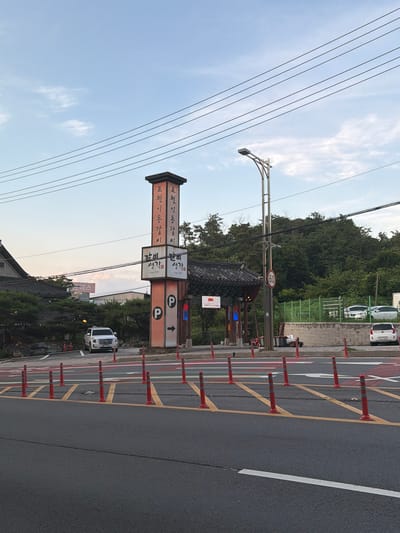 The image shows a street scene in Pocheon-si, South Korea. A tall, orange and white sign is prominently displayed on the right side of the road, featuring Korean text and parking symbols. The sign appears to advertise a restaurant, with the text "포천 이동갈비" (Pocheon Idong Galbi) and "생각" (Saenggak) visible, along with "Since 1997". Next to the tall sign, a smaller sign displays a speed limit of "60". A white SUV is parked in the background, near a building with a traditional Korean roof. Several orange traffic cones are lined up on the side of the road, delineating parking spaces or a traffic lane. The sky is partly cloudy, and green trees and foliage are visible in the background. The overall scene suggests a commercial area, likely a roadside restaurant or a tourist destination.