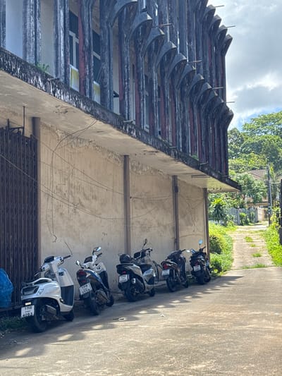 The image depicts the exterior of a weathered building in Ranong, Thailand, characterized by a distinctive facade of vertical, arched concrete structures. A row of parked motorcycles is visible in front of the building. The building's walls show signs of age and disrepair, with peeling paint and a network of overhead wires. To the right of the building, a narrow, unpaved pathway lined with lush green vegetation recedes into the distance, suggesting a residential or less developed area of the city. The sky is partly cloudy.