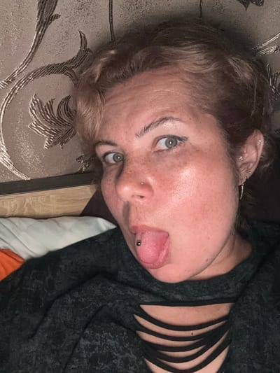 The image is a close-up, low-angle selfie of a person lying down, with their face filling most of the frame. The person has fair skin with freckles, light brown hair styled in soft waves, and green eyes. They are wearing a dark, long-sleeved top with a distinctive cut-out detail across the chest. The person has a tongue piercing with a silver barbell and a small nose stud. Their left ear is visible and adorned with multiple small stud earrings.
The background consists of a textured wallpaper with a subtle brown floral pattern. To the left of the person's head, a portion of a wooden headboard and bedding, including an orange pillow and a white pillow, are visible. The lighting is dim, suggesting an indoor setting, possibly a bedroom. The image does not provide any information that would allow for a specific location in the city of Kozmodemyansk, Russia to be identified.