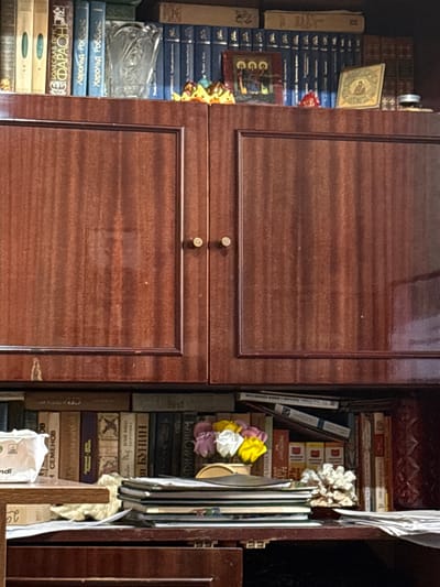 This is a close-up, eye-level shot of a wooden bookshelf filled with books and some decorative items. The bookshelf has a dark, reddish-brown wood finish with a visible grain pattern. The left side of the image shows a cabinet door with two small, round knobs. The right side reveals a section of the bookshelf with books stacked vertically and horizontally.

The books appear to be primarily in Russian, as indicated by the Cyrillic text visible on the spines. Some titles that can be partially deciphered include "Харольд Роббинс" (Harold Robbins) and "АН БУНИН" (A. N. Bunin). There are also some books with simpler colored spines, some blue, some tan, and some brown.

Among the books, there are decorative elements. On the left side, towards the top, is a small, square, framed image with a religious icon. Further down, there's a transparent, possibly glass, decorative object. On the right side, near the middle of the stack of books, are several colorful artificial flowers in shades of yellow, pink, and white.

The lighting is somewhat dim, suggesting an indoor setting, likely a home or library. There are no people visible in the image. The overall impression is one of a well-used personal library, with a mix of literature and sentimental or decorative items. The image is sharp, allowing for clear visibility of the textures of the wood and paper.