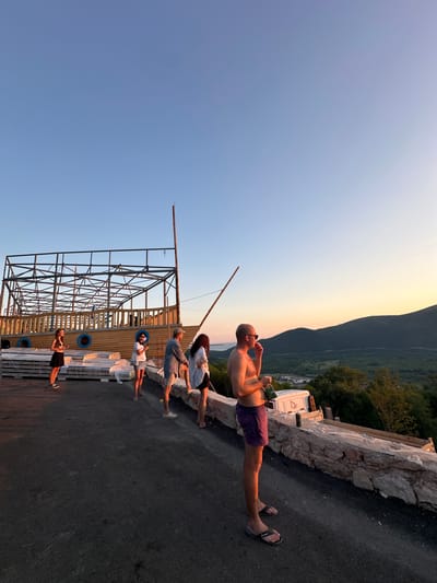 In Svinjišta, Montenegro, at sunset, a group of five people stand near a wooden structure resembling a ship's hull. The structure is under construction, with exposed metal framing above. The sky is clear, transitioning from blue to a warm orange hue near the horizon, with distant mountains silhouetted against it. One man, shirtless and wearing purple shorts, stands in the foreground, facing right and holding a drink. To his left, a man and two women stand, looking towards the scenery. A woman in a black dress stands further left, near stacked materials covered in plastic. The ground is paved, and a low stone wall runs along the edge of the elevated area.