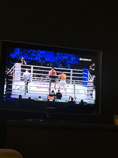 The image shows a television screen displaying a boxing match. Two boxers are in the ring, surrounded by a crowd. The ring has advertisements for "ELITBET," "GymBeam," and "PROPER TWELVE." Text on the screen shows the names of the boxers as "S. KUNOV" and "C. MALICHOUDIS." A logo reads "ACTION." The television is a Samsung. It is not possible to determine if the scene is from the city of Ede, Netherlands, or to infer any location or connection.