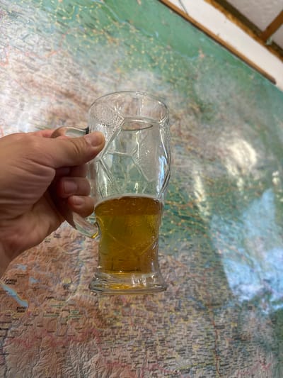 A person's hand holds a glass mug filled with beer in front of a map. The map appears to be of Bulgaria, with some place names visible in Bulgarian script, including "Минерални Бани" (Mineralni Bani). The beer in the mug is a golden amber color with a frothy head. The glass mug has a textured pattern. The background is a wall with a large map mounted on it, and part of a ceiling is visible at the top. The lighting suggests an indoor environment.