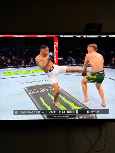 The image shows a television screen displaying a UFC (Ultimate Fighting Championship) match. The screen shows two fighters engaged in combat. One fighter is wearing white shorts, while the other is wearing green and yellow shorts. The screen also displays the names of the fighters as "1 ДЕЛЛА МАДДАЛЕНА" and "5 ПРАТЕС". The UFC logo and a timer showing "1:14" are visible, along with the text "ПОЛУСРЕДНИЙ ВЕС". Advertisements for Budweiser, Ladbrokes, Spudshed, and Monster Energy are displayed. The image does not show the city of Ligachevo, Russia.